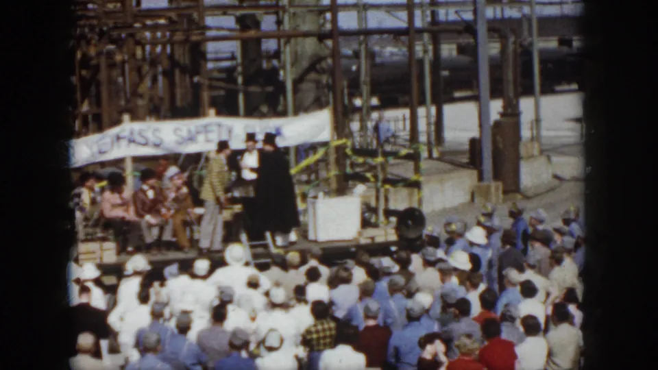 1958 Virginia frame of a public gathering at an industrial site, with speakers on stage and a safety-show banner behind them.