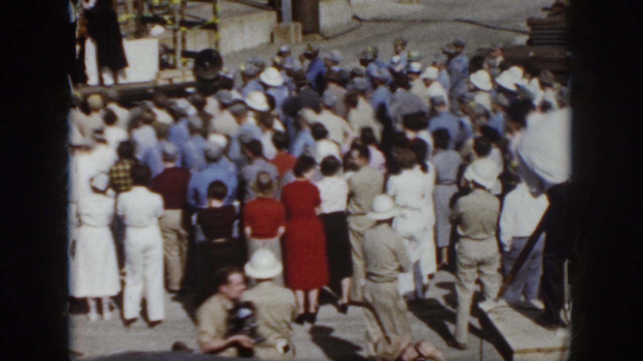 Crowd in hard hats and 1950s clothing gathered near an industrial stage.