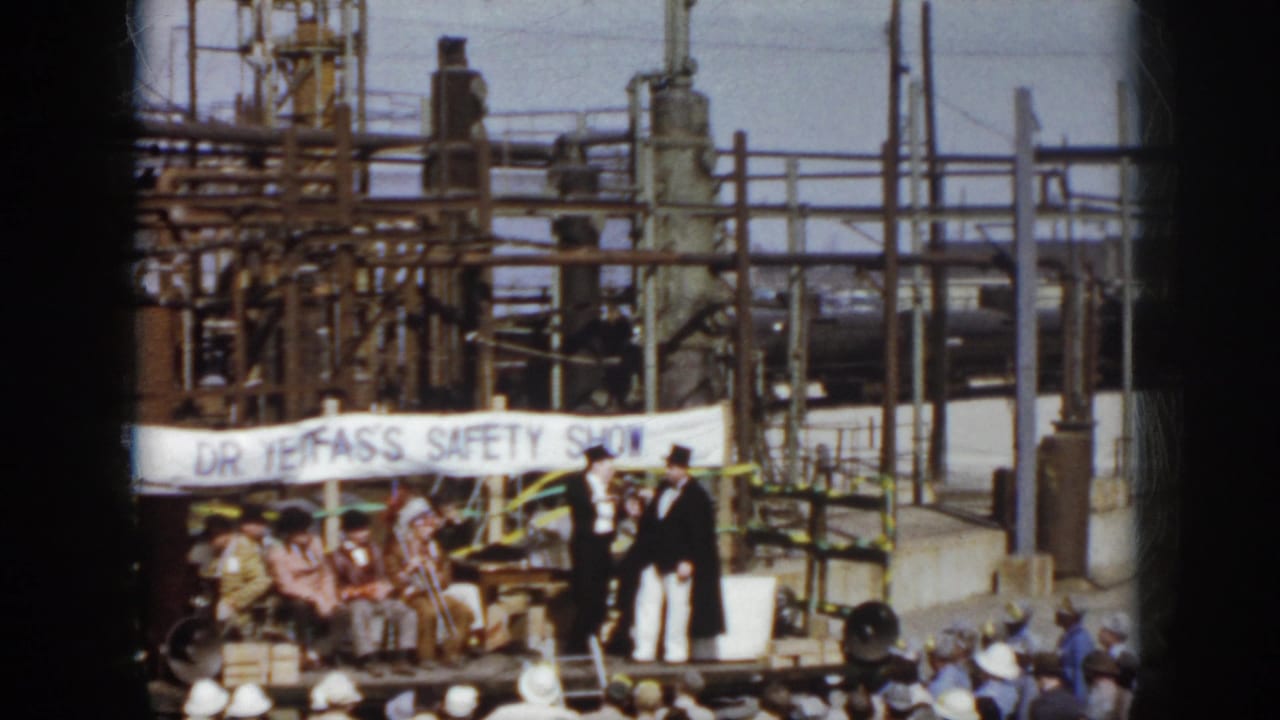 1958 Virginia home-movie frame showing a small outdoor safety-show stage, performers in formal costumes, an audience, and industrial pipework behind them.