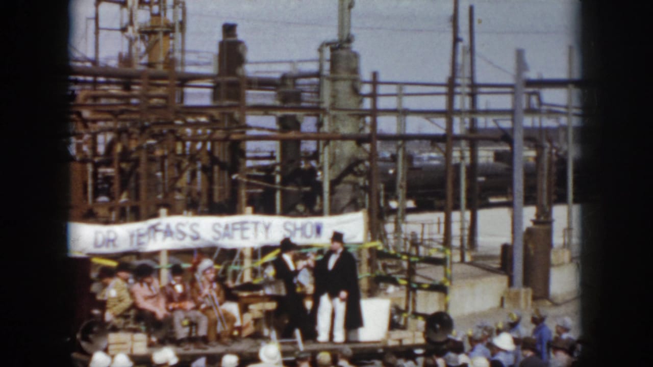 Two formally dressed men perform on a stage under a banner, with industrial structures in the background.
