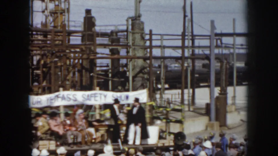 1958 Virginia home-movie view of seated spectators and formally dressed performers on an outdoor safety-show stage at an industrial site.