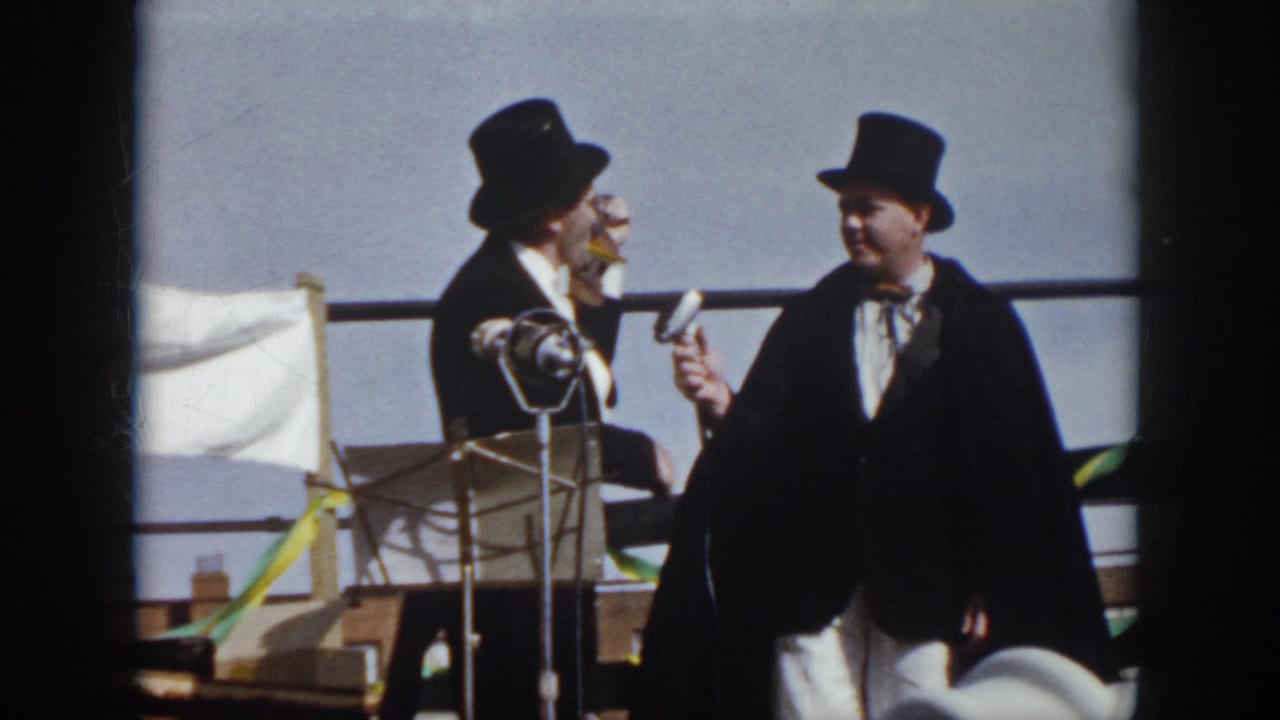 1958 Virginia 8mm frame showing two formally dressed men in top hats on an outdoor stage with microphones, a white flag, and streamers.