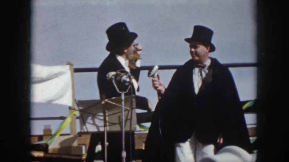 1958 Virginia 8mm frame showing two formally dressed men in top hats on an outdoor stage with microphones, a white flag, and streamers.