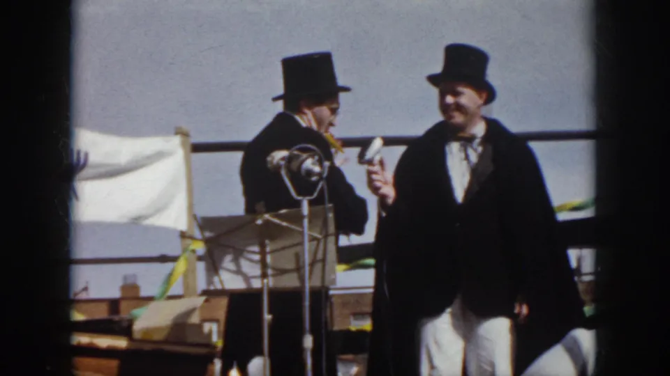 1958 Virginia home-movie frame with one performer holding a microphone toward another man in a top hat near a standing microphone.