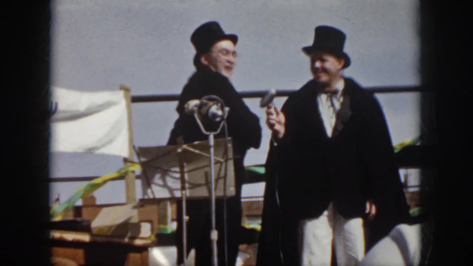 1958 Virginia 8mm frame showing two men in formal stage costume facing outward near microphones, with a flag and streamers in the background.