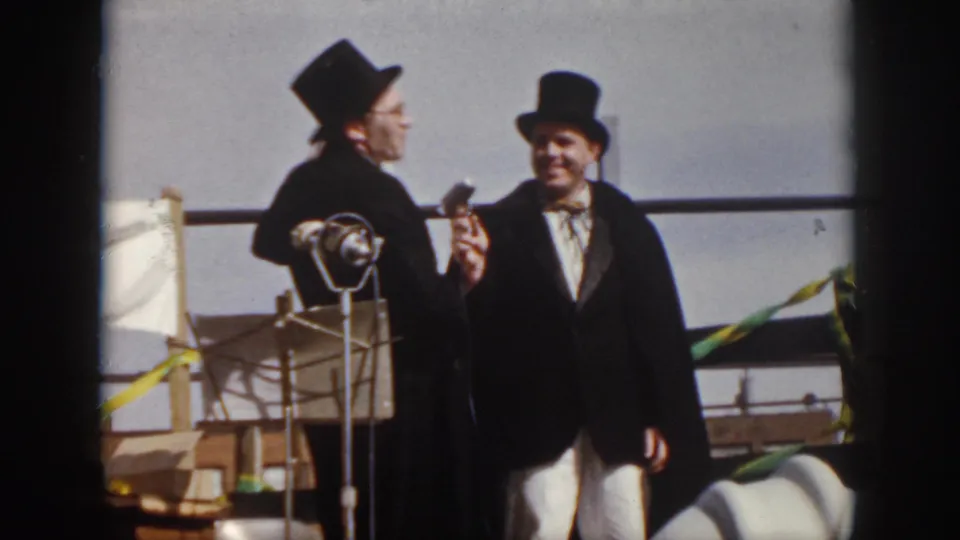 1958 Virginia home-movie frame with two smiling performers in top hats and capes standing near microphone stands on an outdoor stage.