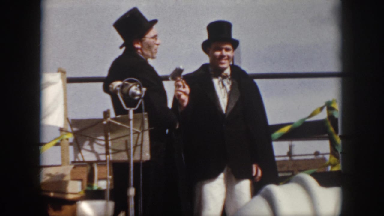 Two men in top hats stand on an outdoor stage, one near a microphone.