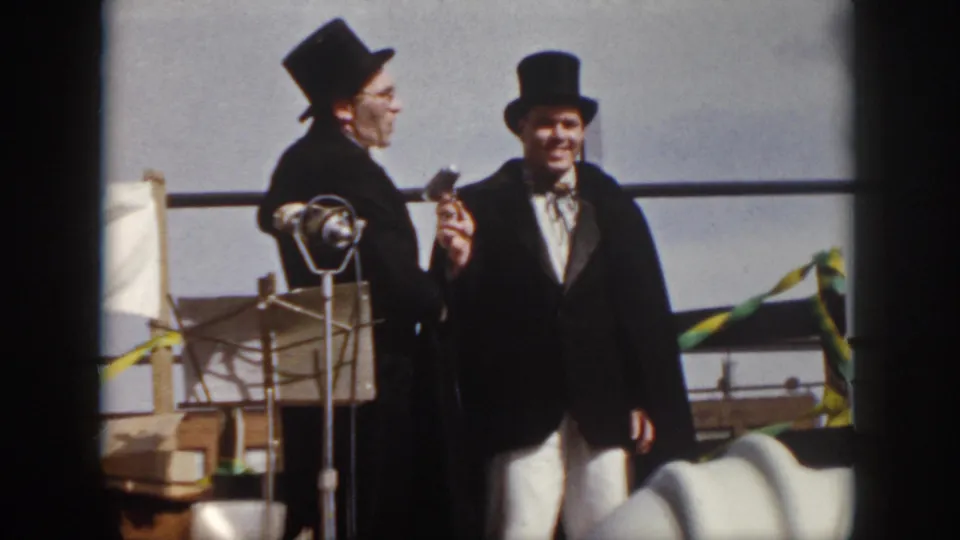 1958 Virginia home-movie frame showing two formally dressed men in top hats on a small outdoor stage with a microphone.