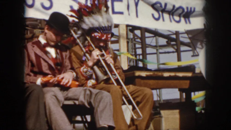 1958 Virginia frame of seated stage musicians, including a violin player and a trombone player wearing a feathered headdress.