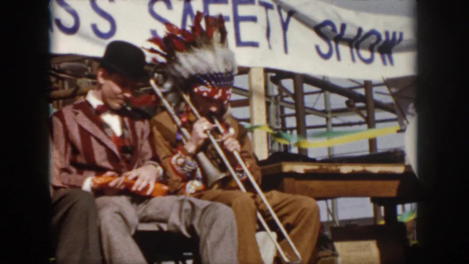 1958 Virginia home-movie frame with a bowler-hatted violinist seated beside a trombone player in a Native American-style headdress.
