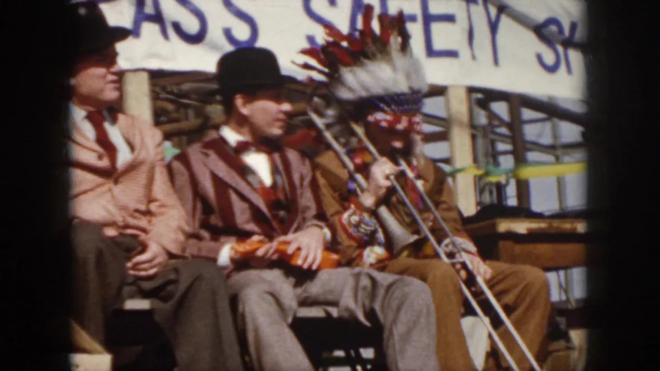 1958 Virginia 8mm frame showing men seated on stage in suits and bowler hats beside a trombone player in a Native American-style headdress.