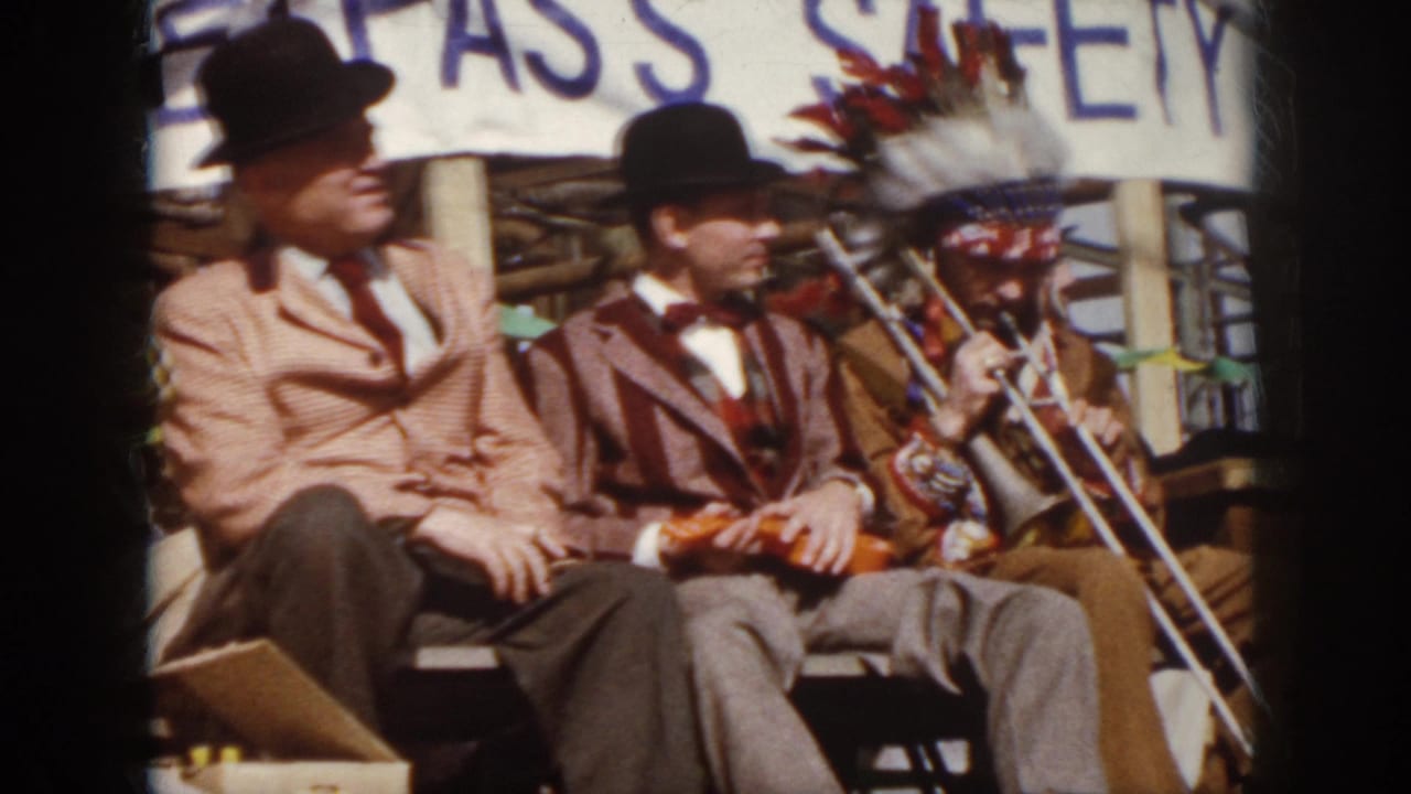 1958 Virginia home-movie frame of seated stage performers in suits and bowler hats, with a trombone and headdress visible at right.