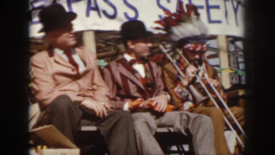 1958 Virginia home-movie frame of seated stage performers in suits and bowler hats, with a trombone and headdress visible at right.