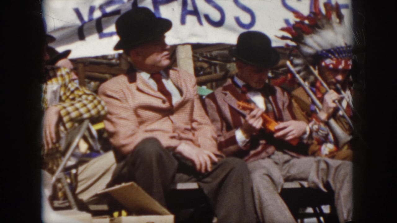 1958 Virginia 8mm frame with suited men seated on stage, a trombone player in headdress nearby, and a plaid-costumed figure partly visible.
