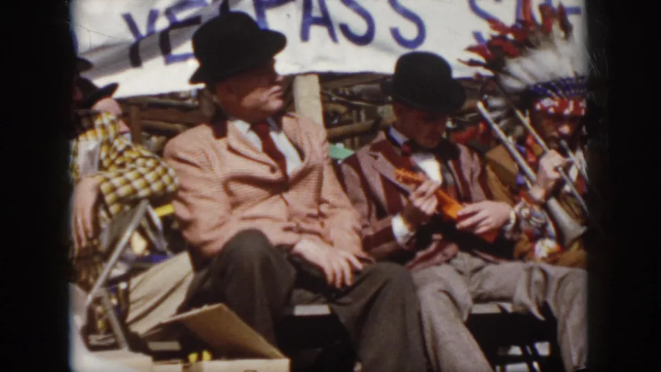 1958 Virginia 8mm frame with suited men seated on stage, a trombone player in headdress nearby, and a plaid-costumed figure partly visible.