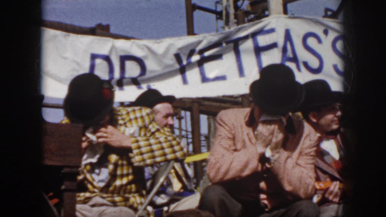 1958 Virginia home-movie frame showing seated costumed musicians under a banner, with one performer playing harmonica.