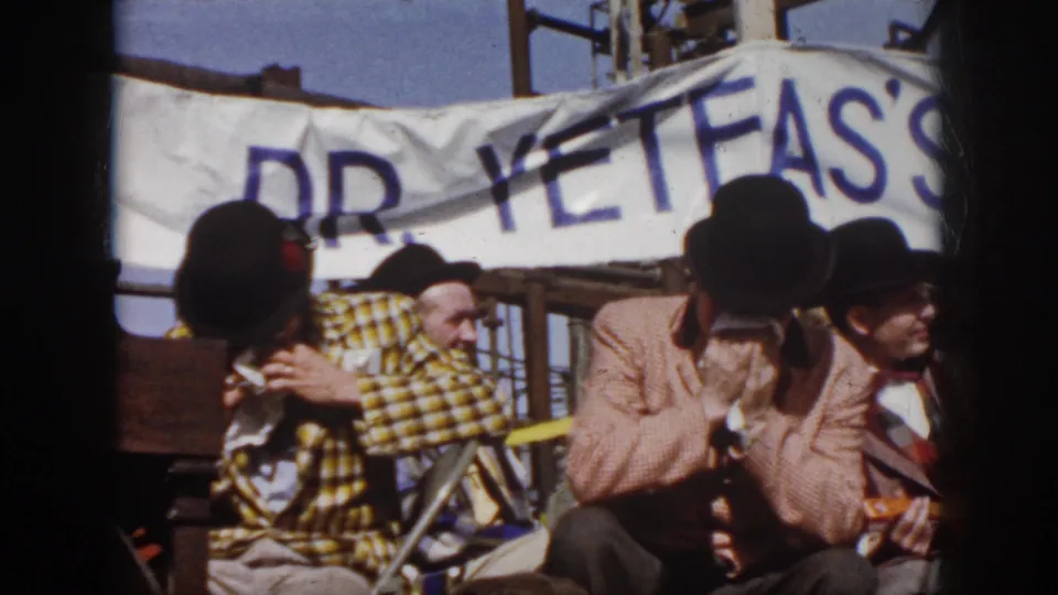 1958 Virginia home-movie frame showing seated costumed musicians under a banner, with one performer playing harmonica.