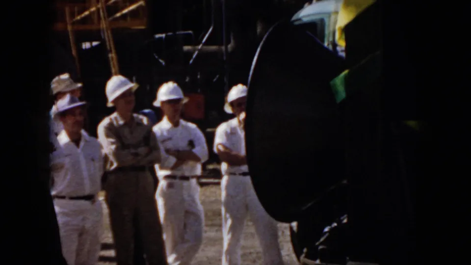 1958 Virginia factory or plant scene with men in hard hats and work uniforms listening near large black speakers.