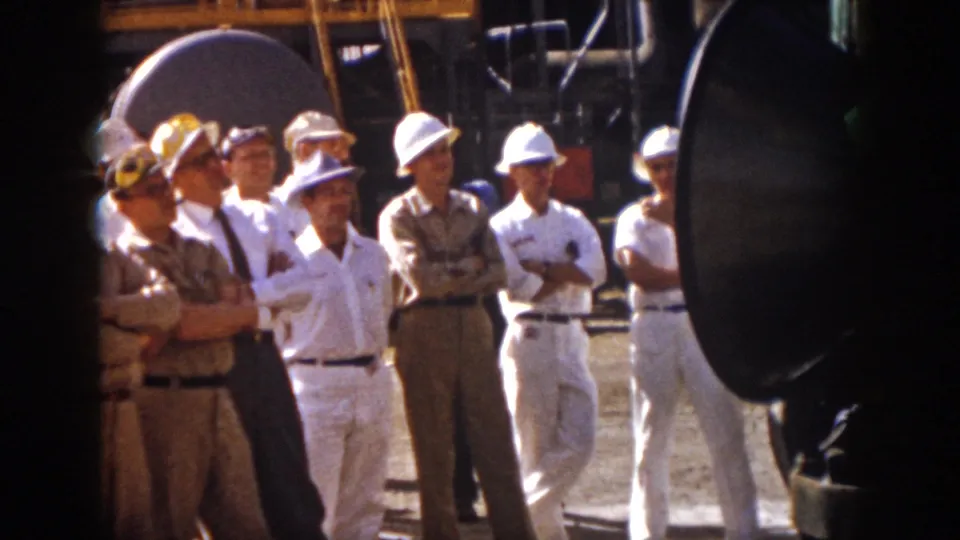 Men in white hard hats, caps, white coveralls, and khaki uniforms listen in a Virginia industrial setting in 1958.