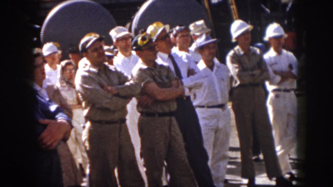 1958 Virginia home-movie frame of men in work uniforms and hard hats listening beside large industrial loudspeakers.