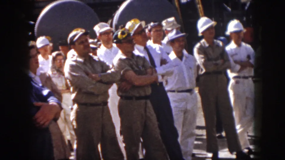 1958 Virginia home-movie frame of men in work uniforms and hard hats listening beside large industrial loudspeakers.