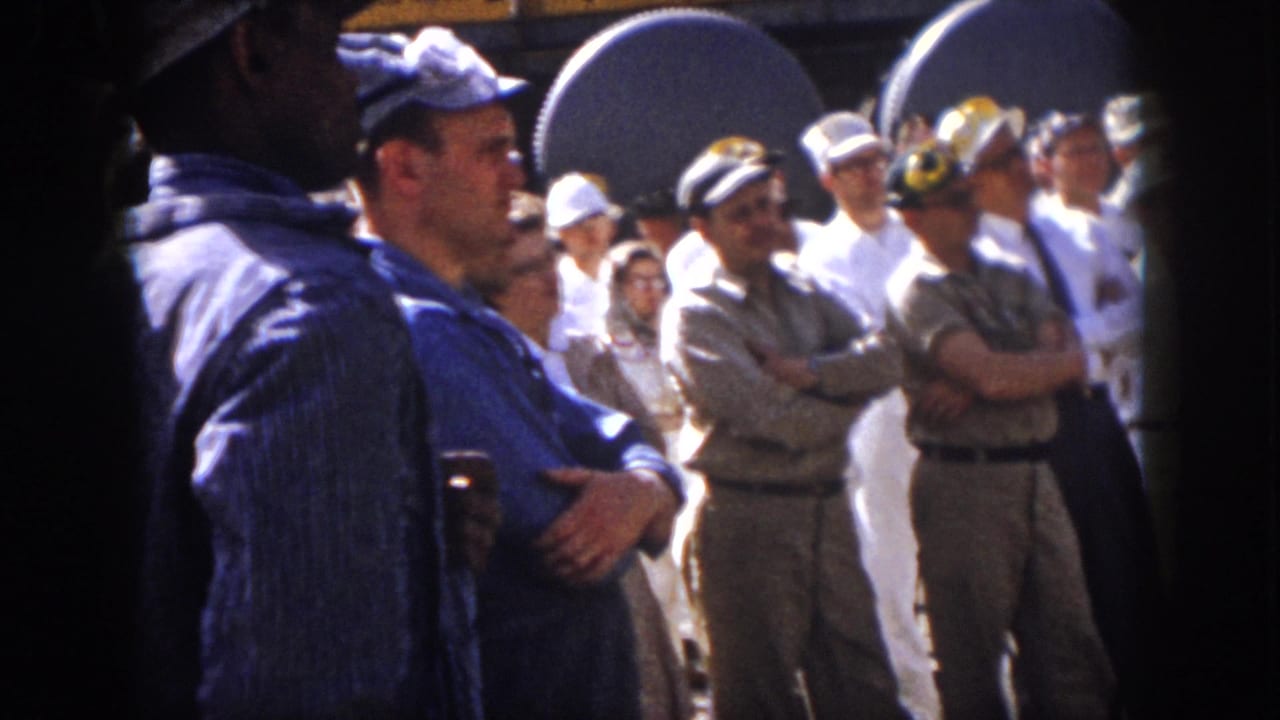 1958 Virginia frame with uniformed men, hard hats, and visitors watching something off-camera at an industrial site.