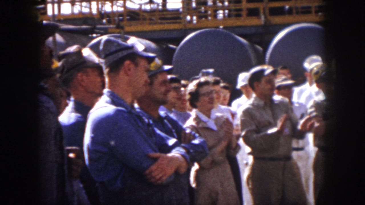 1958 Virginia home-movie frame showing workers in caps, bystanders, and a woman among the crowd during an outdoor industrial-site gathering.