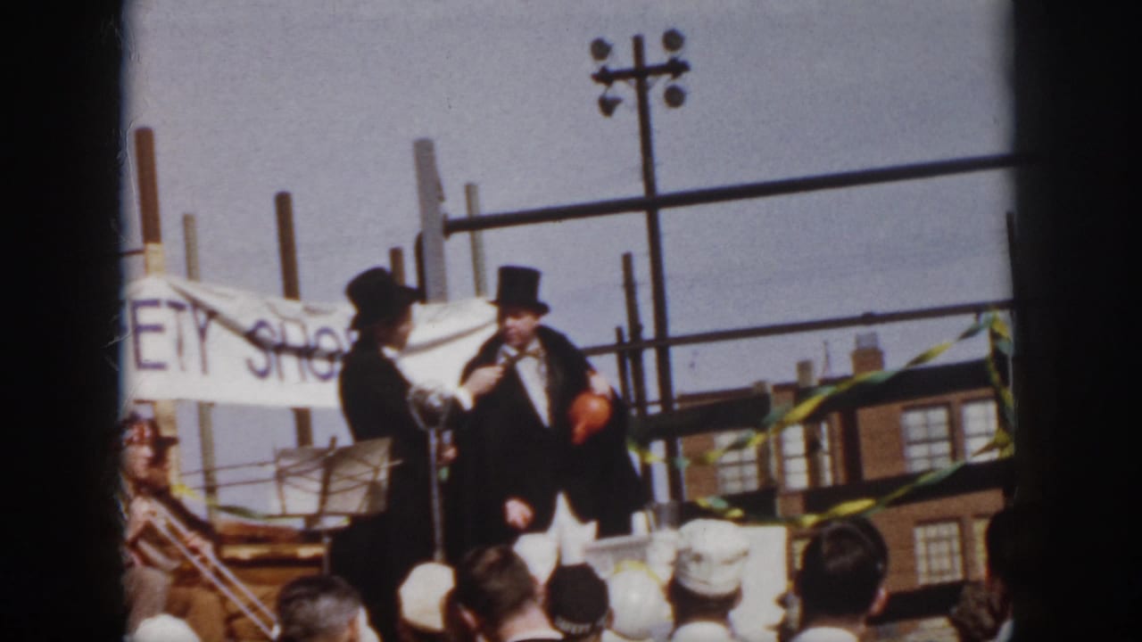 Two men in top hats and dark suits stand on an outdoor stage before a crowd.