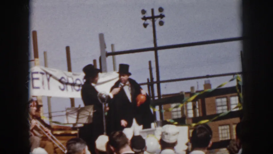 1958 Virginia frame of two formally dressed men on a makeshift outdoor platform with a microphone, bunting, and a partial banner behind them.