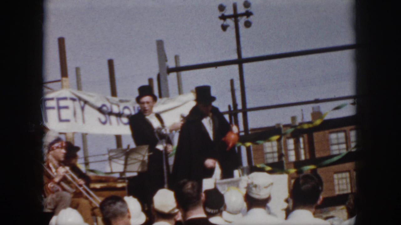 Mid-century Virginia home-movie frame showing suited men on an outdoor platform, with spectators, bunting, and a partly visible banner behind them.