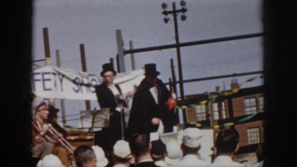 Mid-century Virginia home-movie frame showing suited men on an outdoor platform, with spectators, bunting, and a partly visible banner behind them.
