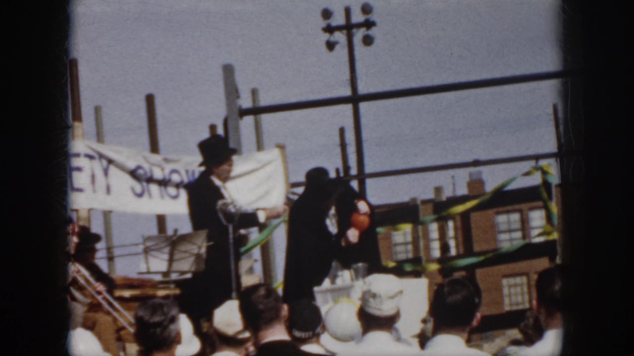 1958 Virginia outdoor gathering frame with top-hatted presenters on stage, striped bunting, utility poles, and brick buildings in the background.