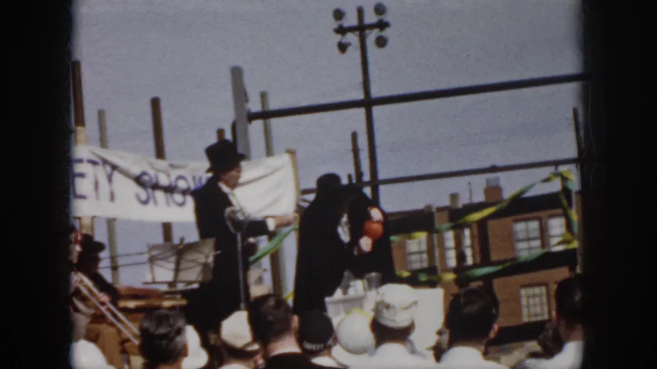 1958 Virginia outdoor gathering frame with top-hatted presenters on stage, striped bunting, utility poles, and brick buildings in the background.
