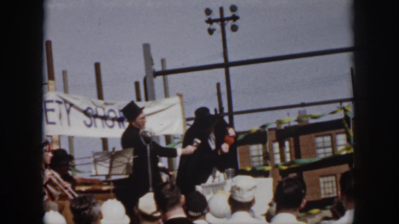 In Virginia, 1958, two costumed male performers stand near a microphone on a makeshift outdoor stage as spectators watch.
