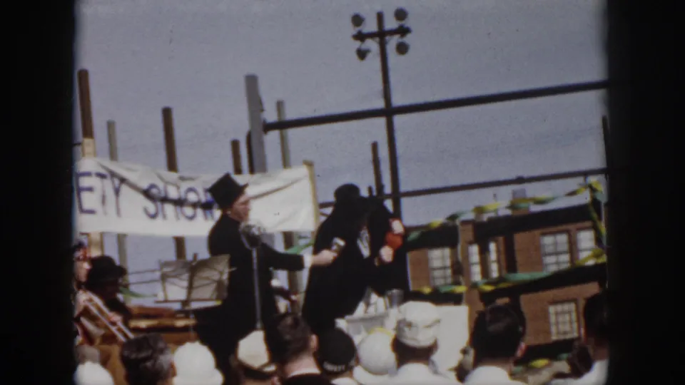 In Virginia, 1958, two costumed male performers stand near a microphone on a makeshift outdoor stage as spectators watch.