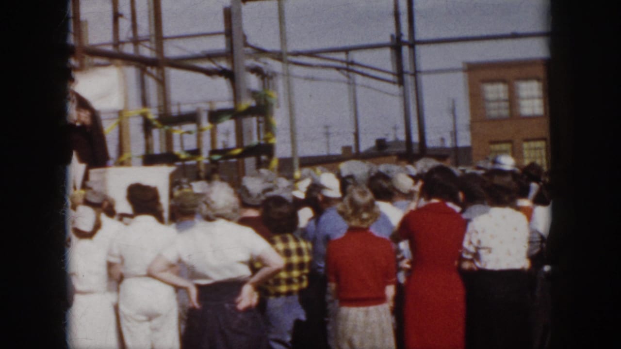 Spectators in 1950s clothing stand with their backs to the camera while a distant outdoor stage sits among poles and brick buildings in Virginia.