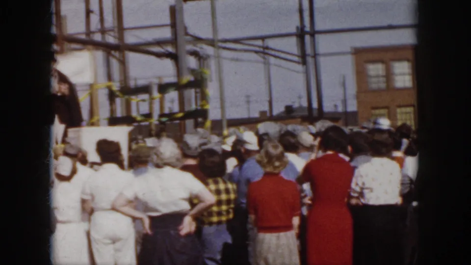 Spectators in 1950s clothing stand with their backs to the camera while a distant outdoor stage sits among poles and brick buildings in Virginia.