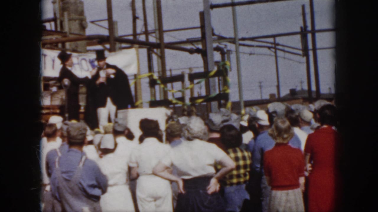 1958 Virginia home-movie frame showing spectators facing two formally dressed men on a makeshift industrial stage with safety signage.