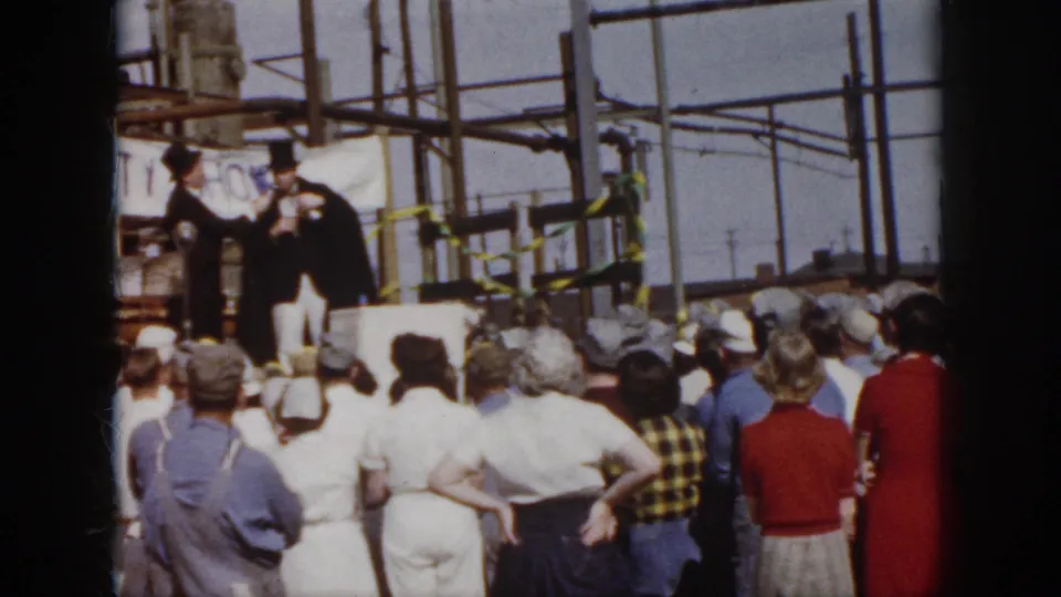 1958 Virginia home-movie frame showing spectators facing two formally dressed men on a makeshift industrial stage with safety signage.