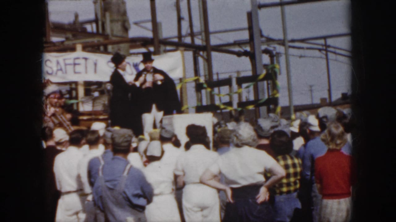 1958 Virginia footage frame with a crowd of workers and spectators watching two top-hatted speakers near a visible safety banner.