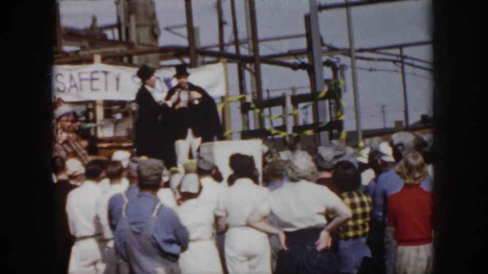 1958 Virginia footage frame with a crowd of workers and spectators watching two top-hatted speakers near a visible safety banner.