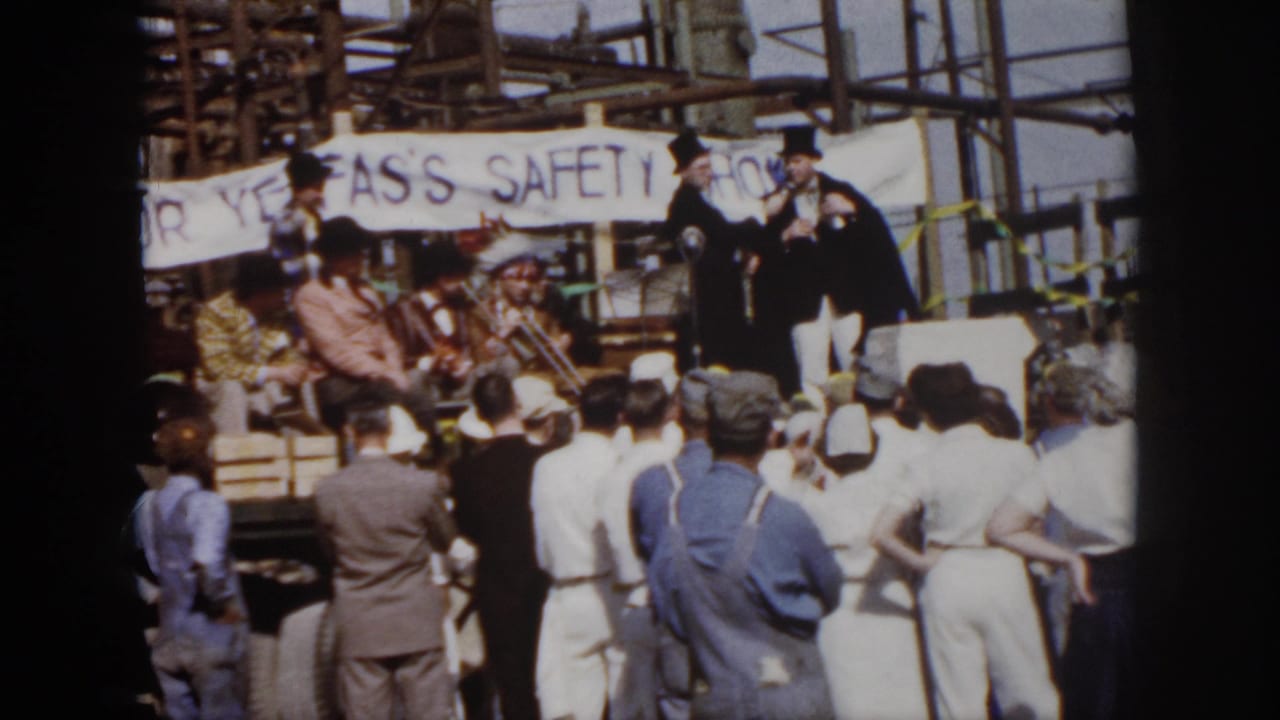 1958 Virginia home-movie frame showing spectators facing a small outdoor stage with top-hatted performers and a safety-show banner.