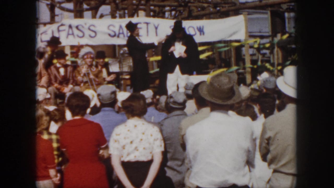 1958 Virginia footage frame with a rear view of spectators watching formal-costumed performers on an outdoor safety-show stage.