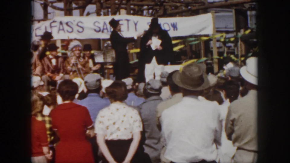 1958 Virginia footage frame with a rear view of spectators watching formal-costumed performers on an outdoor safety-show stage.