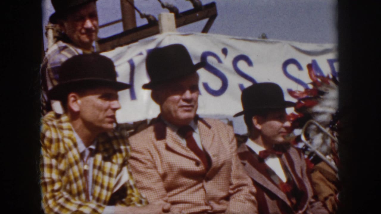 1958 Virginia home-movie frame showing men in bowler hats and suits seated on a parade float with a safety banner behind them.