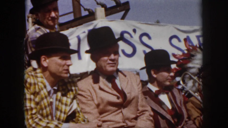 1958 Virginia home-movie frame showing men in bowler hats and suits seated on a parade float with a safety banner behind them.