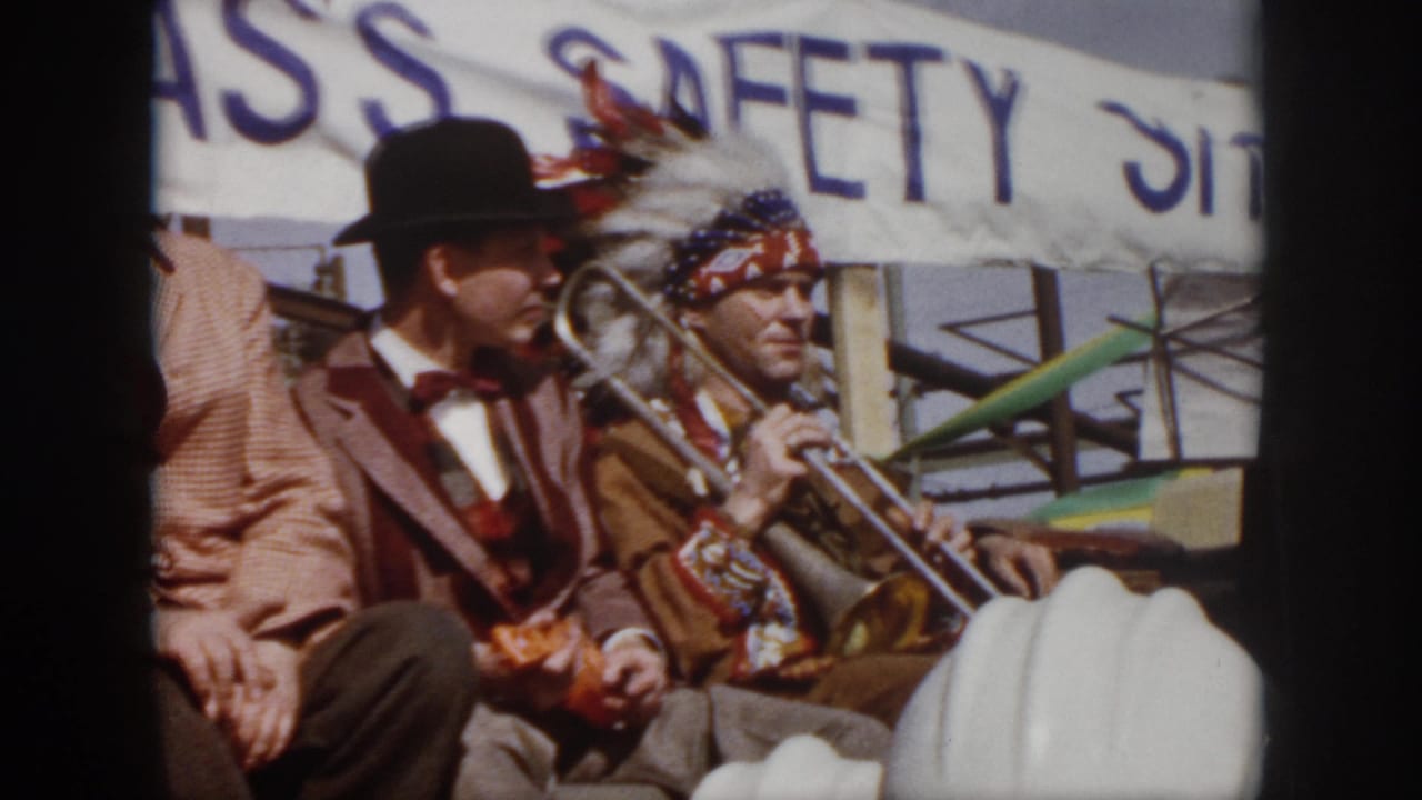 1958 Virginia parade frame showing a costumed trombone player and suited men seated on a float beneath a safety banner.