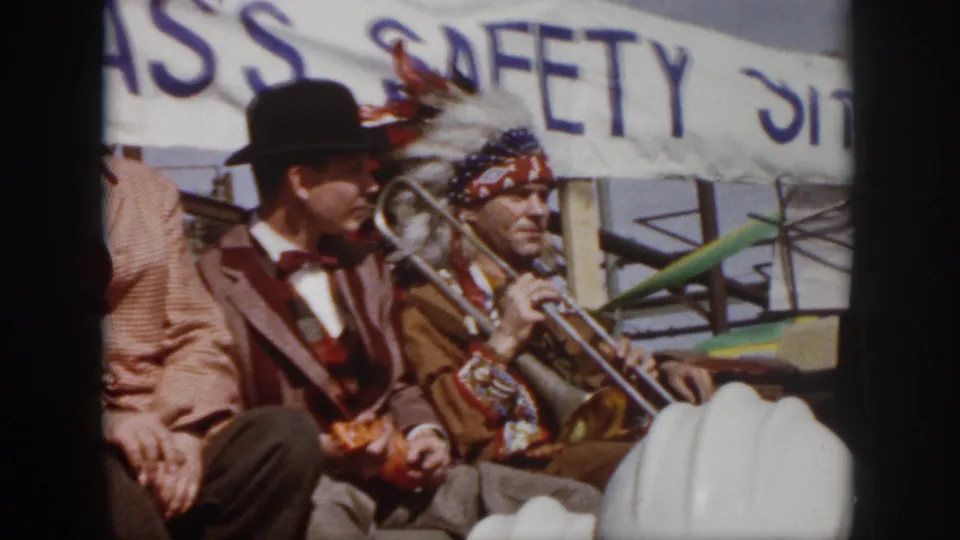 1958 Virginia parade frame showing a costumed trombone player and suited men seated on a float beneath a safety banner.