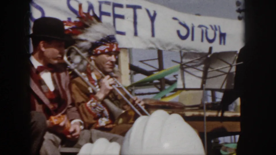1958 Virginia workplace safety event frame with performers seated onstage, a readable SAFETY SHOW banner, and hard hats in the audience foreground.