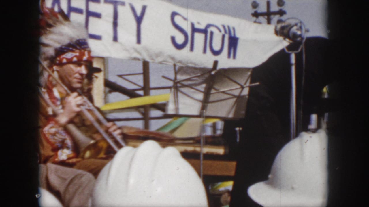 1958 Virginia 8mm frame showing a safety show performer at left, white hard hats in the foreground, and a partial suited figure crossing the stage area.